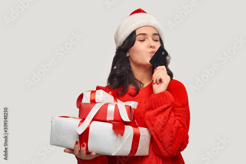 Girl in a red sweater and Santa hat holding giftboxes for Christmas and kissing bank card