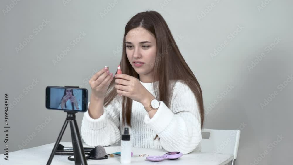 grey back Caucasian girl blogger white jumper Straight long hair Desk in front of cellphone unscrews tube of liquid lipstick demonstrates. On table are brushes Screen phone deployed on viewer side grey back Caucasian girl blogger white jumper Straight long hair Desk in front of cellphone unscrews tube of liquid lipstick demonstrates. On table are brushes Screen phone deployed on viewer side
