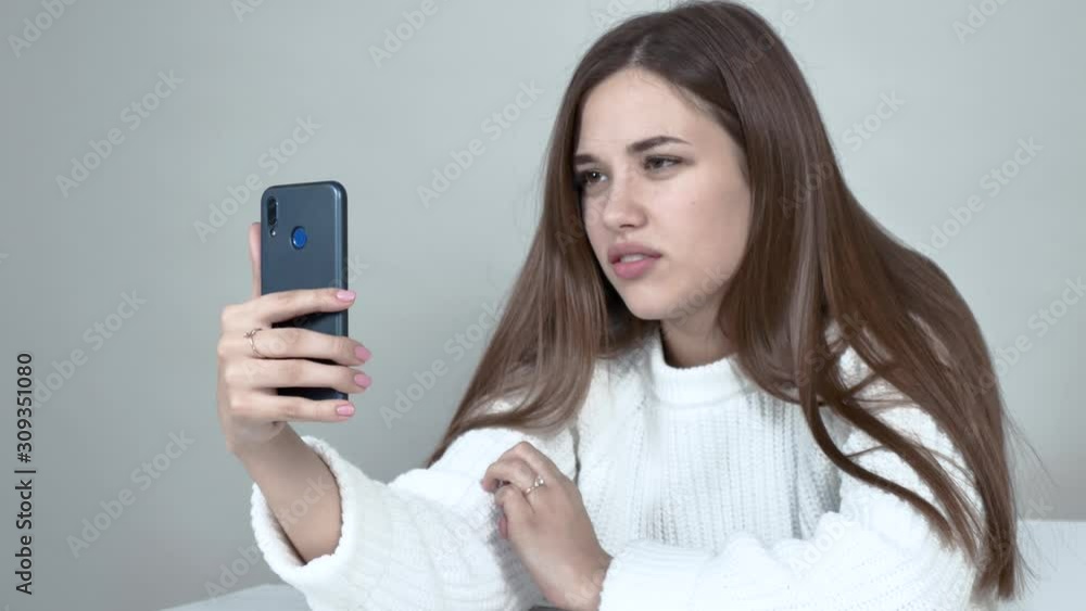Grey background Caucasian girl white jumper. Her hair is straight and falls over her shoulders. She sits at a table, holding smart phone looking at camera, frowns, something Neubeuern says. Close up