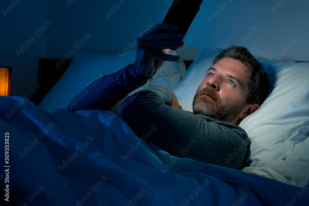 Fototapeta premium young attractive and relaxed man with blue eyes lying on bed late at night in dark and dim light networking on mobile phone or online dating in internet addiction