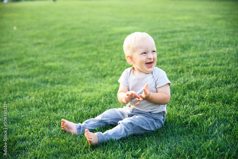 Fototapeta premium Little boy with blond hair laughs sitting on the green grass in the park