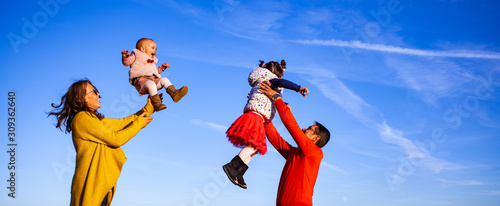 happy family of four mother and father throwing kids up against blue sky panoramic shot