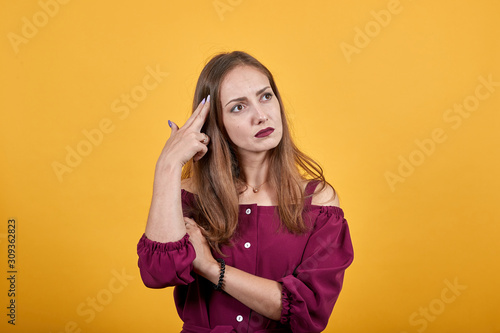Ginger girl in burgundy bluse with bow over isolated orange background thoughtfully looks right holding two fingers on temple