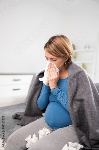 Pregnant woman catching cold, flu, virus, sitting at home on a couch, blowing nose and beaing sick.