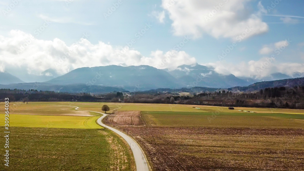 A panoramic, drone shot of an alpine landscape of Austria. Lush green meadows and crop fields spread on a vast surface. Idyllic landscape. There is a road leading to the mountains in the middle.