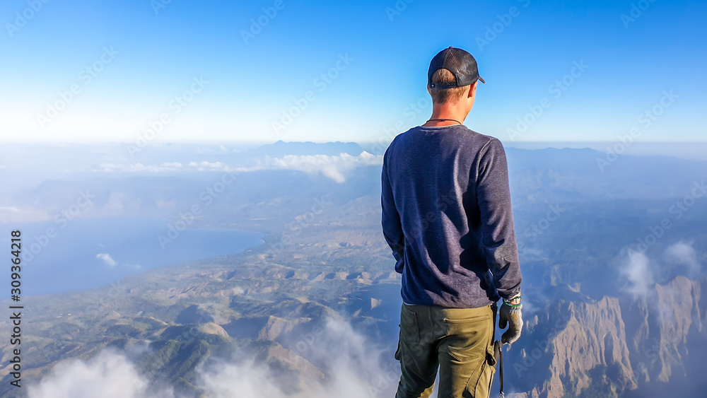 A young man standing of the top of volcano Inerie in Bajawa, Flores, Indonesia. He is enjoying the beautiful view on volcanic island. There are some clouds below him. Discovering while travelling.