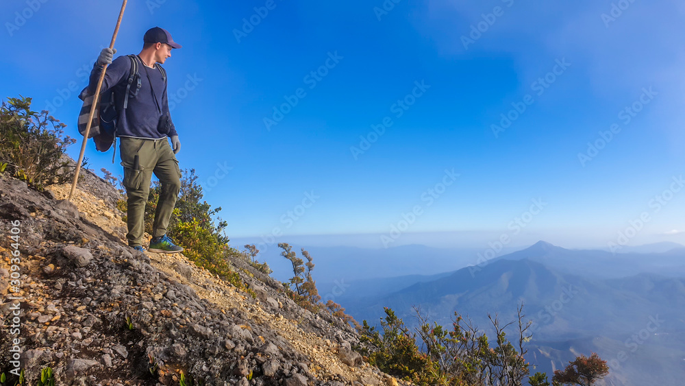A young man walking down the volcano Inerie in Bajawa, Flores, Indonesia. He supports himself on a wooden stick while enjoying the beautiful view on volcanic island. Serenity and calmness.