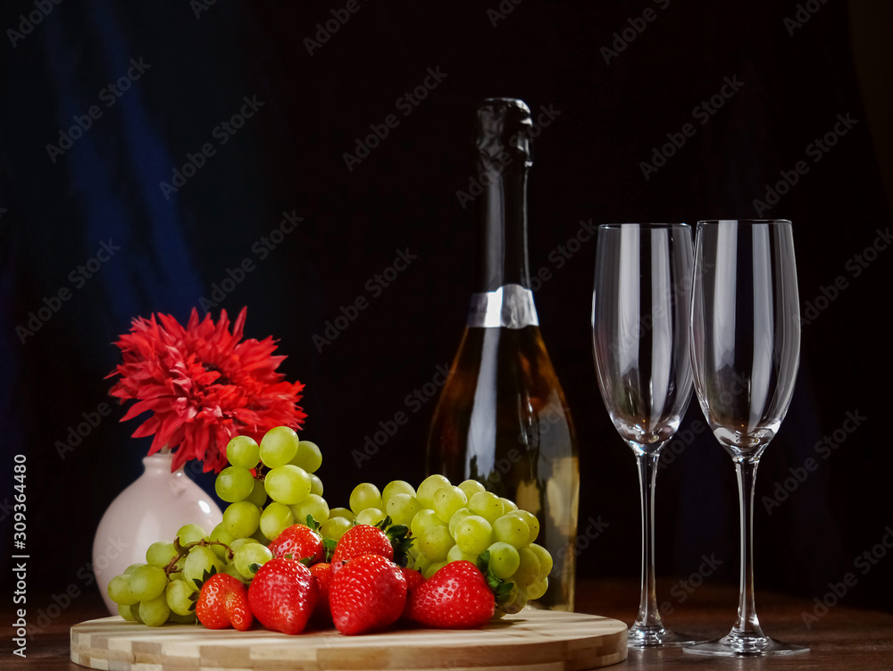 Fresh green grapes and red strawberry on a wooden board, Two champagne flutes  and a bottle of champagne without labell, flower in the background, Dark background. Selective focus.