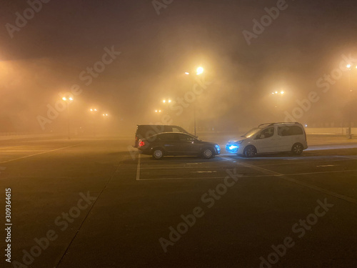Kehl  Germany - Dec 04  2019  Tight fog on the large parking illuminated by multiple lights and three parked cars
