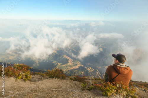 Fototapeta Naklejka Na Ścianę i Meble -  A young man sitting of the top of volcano Inerie in Bajawa, Flores, Indonesia. He is enjoying the beautiful view on volcanic island. There are some clouds below him. Discovering while travelling.
