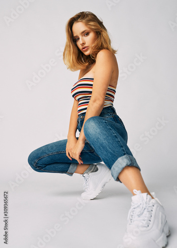 squatting young and athletic girl in jeans and a short T-shirt, beautiful and healthy skin, natural blond hair, empty background, delicate makeup,