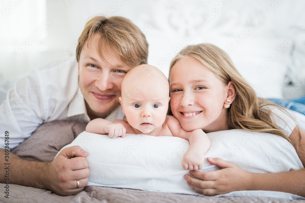 Beautiful smiling young family mom dad elder daughter and newborn baby are lying on a large bed in a bright bedroom. Concept of friendly caucasian family Beautiful smiling young family mom dad elder daughter and newborn baby are lying on a large bed in a bright bedroom. Concept of friendly caucasian family