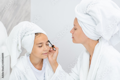 Mother and young daughter applying makeup at home. Mom and child girl are in bathrobes and with towels on their heads