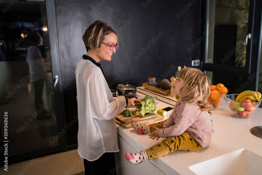 Mother cutting vegetables in the kitchen talking to a little girl sitting next to her