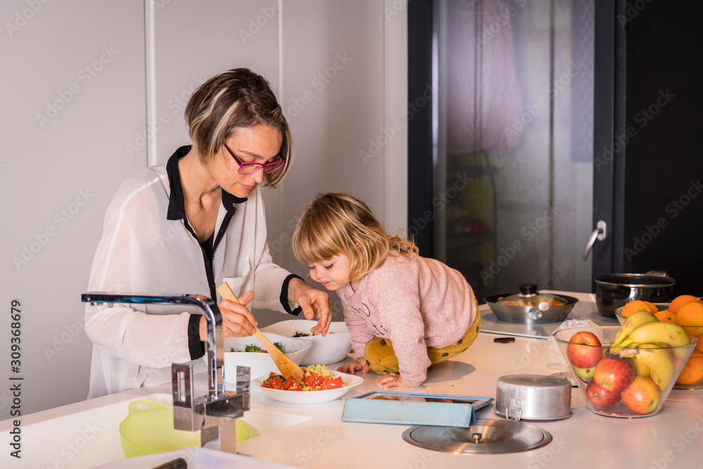 Mother preparing salad in the kitchen with a little girl sitting next to her who helps he