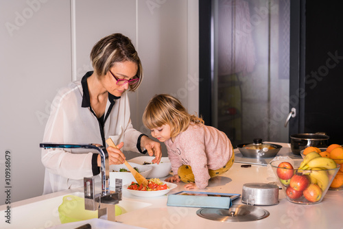 Obraz na płótnie Mother preparing salad in the kitchen with a little girl sitting next to her who helps he w salonie