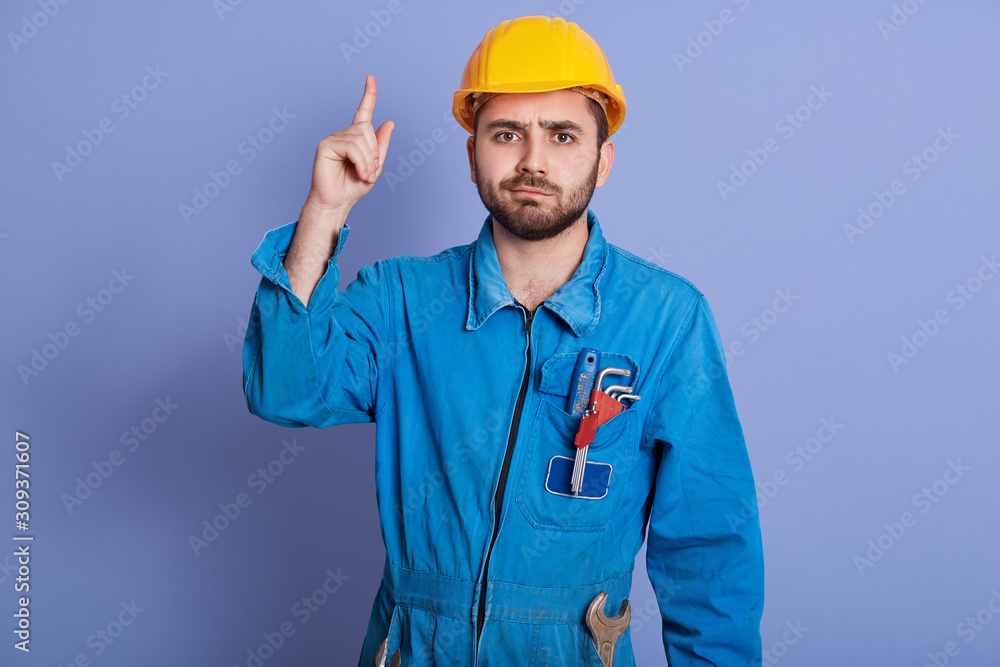 Studio shot of young construction worker with tools in pocket, dresses yellow helmet and uniform pointing up, bearded male having serious expression, copy space for advertisment or promotional text. Studio shot of young construction worker with tools in pocket, dresses yellow helmet and uniform pointing up, bearded male having serious expression, copy space for advertisment or promotional text.