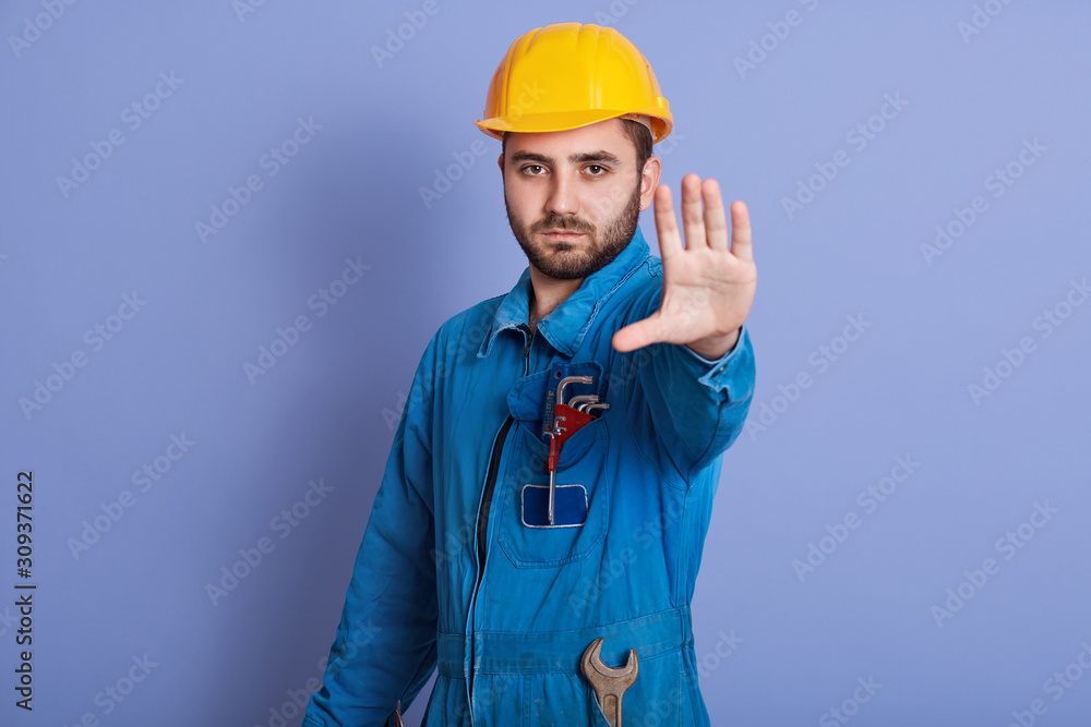 Young bearded handsome workman with yellow helmet and uniform making stop gesture with his hand denying situation that thinks wrong, posing with serious expression isolated over blue background.