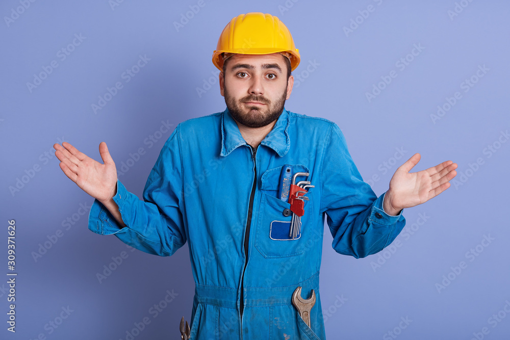 Close up portrait surprised bearded builder dresses helmet and uniform, spreading his hands to sides, squeezing his shoulders, posing isolated over blue background, guy with wrench tools working. Close up portrait surprised bearded builder dresses helmet and uniform, spreading his hands to sides, squeezing his shoulders, posing isolated over blue background, guy with wrench tools working.