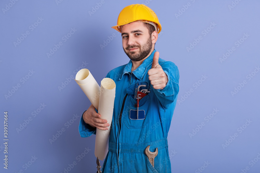 Young beraded caucasian architect with helmet, handsome man posing with thumb up and holding bluepints inhands, wearing blue uniform and yellow hard hat, working on new project, having great succses. Young beraded caucasian architect with helmet, handsome man posing with thumb up and holding bluepints inhands, wearing blue uniform and yellow hard hat, working on new project, having great succses.