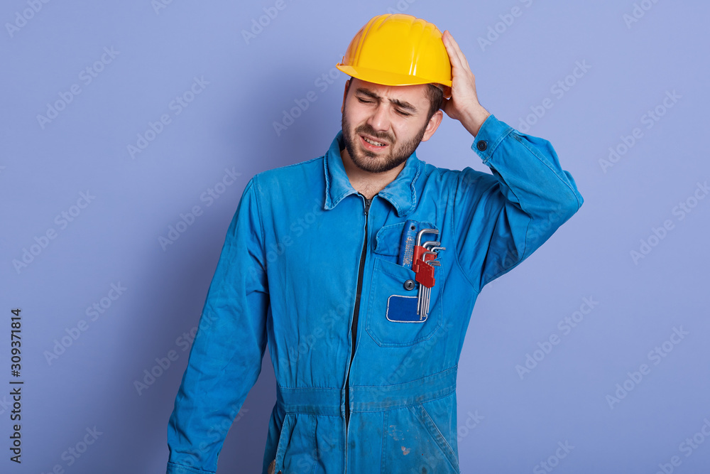 Studio shot of handsome young bearded Caucasian mechanic wearing blue work clothes and yellow hard hat, stands touching his head, having painful look, feeling exhausted after hard working day.