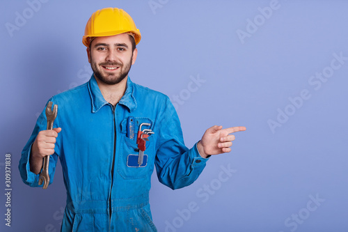 Happy and smiling bearded engineer with wrench tool in hand looking at camera and pointing aside with index finger while stands against blue studio background, dresses blueuniform and yellow helmet. photo