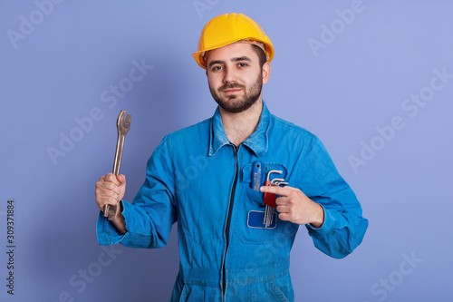Close up portrait of bearded engineer with wrenches looking at camera and pointing athis tool with index finger, repairman posing against blue background, mechanic dresses uniform and yellow hard hat. photo