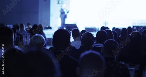 People listening unrcognizable businesswoman talking on stage and showing presentation. Viewers sitting back to camera on business seminar, view from auditorium. Educational conference concept.