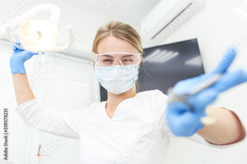 Confident dentist holds her instrument in one hand and move lamp closer. Selective focus