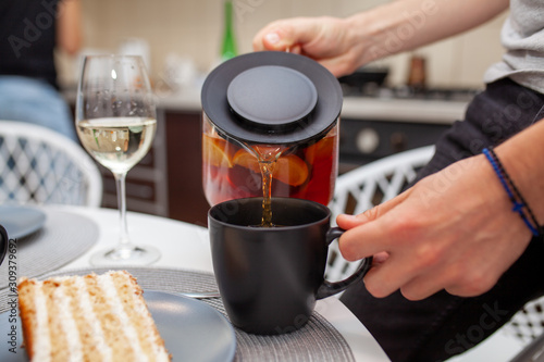 Closeup of male hands pouring tea into a cup. On the side on the table is a glass of wine and a piece of cake