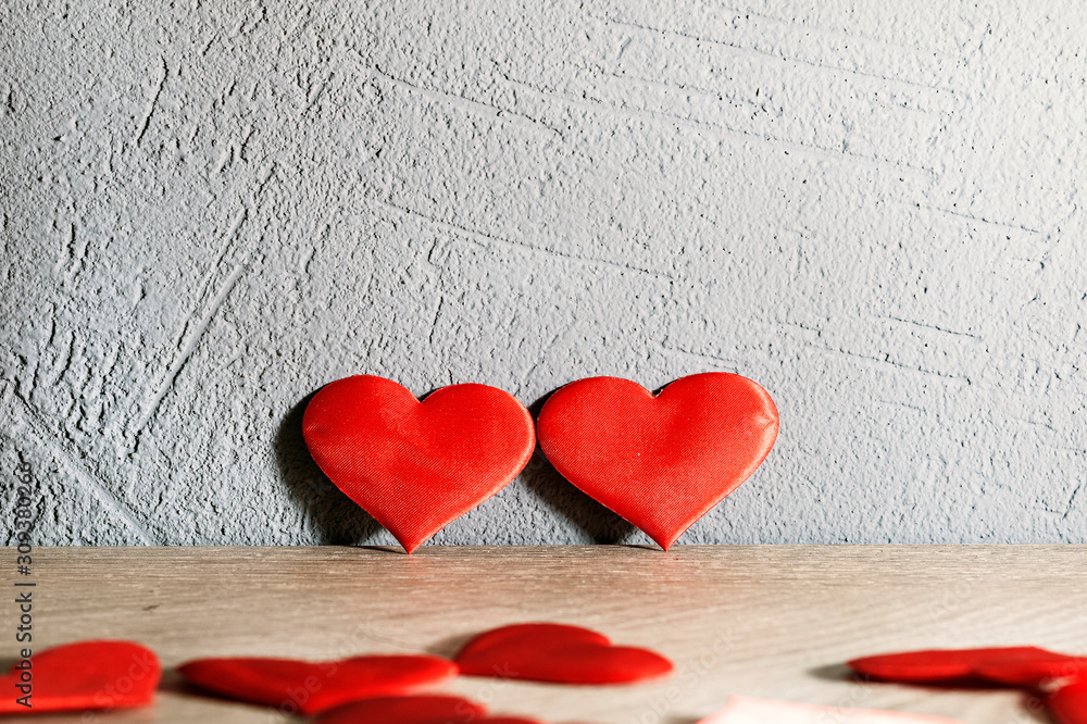 two red hearts stand next to a wooden table against a textured wall. other hearts are scattered across the table.. two red hearts stand next to a wooden table against a textured wall. other hearts are scattered across the table..