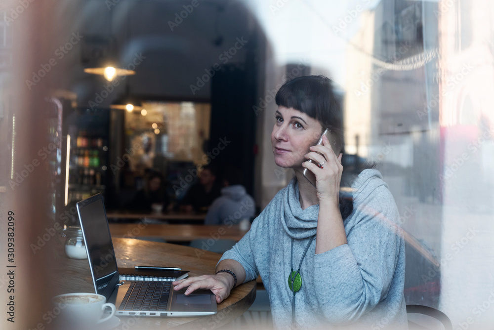 Woman with laptop is taliking by mobile phone and is online, she's working in coworking in cafe as a freelancer. Woman is sitting near the window, there is a reflection in the glass. Woman with laptop is taliking by mobile phone and is online, she's working in coworking in cafe as a freelancer. Woman is sitting near the window, there is a reflection in the glass.