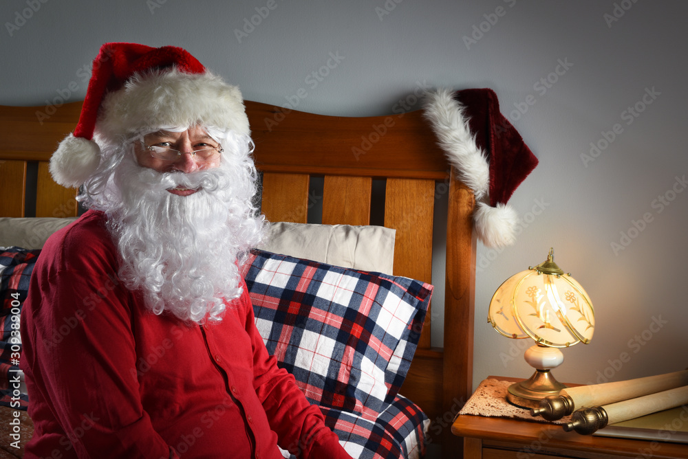 Closeup of Santa Claus wearing a red union suit sitting on the edge of his  bed at the North Pole, ready to go to sleep after delivering packages on  Christmas eve. Stock