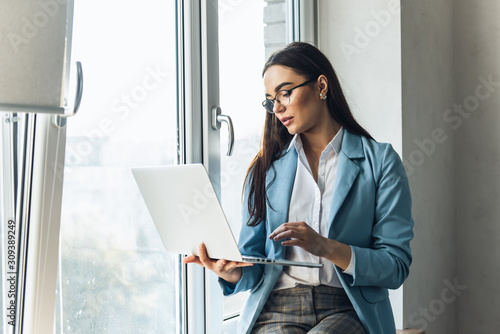 Photography Young beautiful businesswoman in glasses working on laptop and keeping hand on chin while sitting at her working place