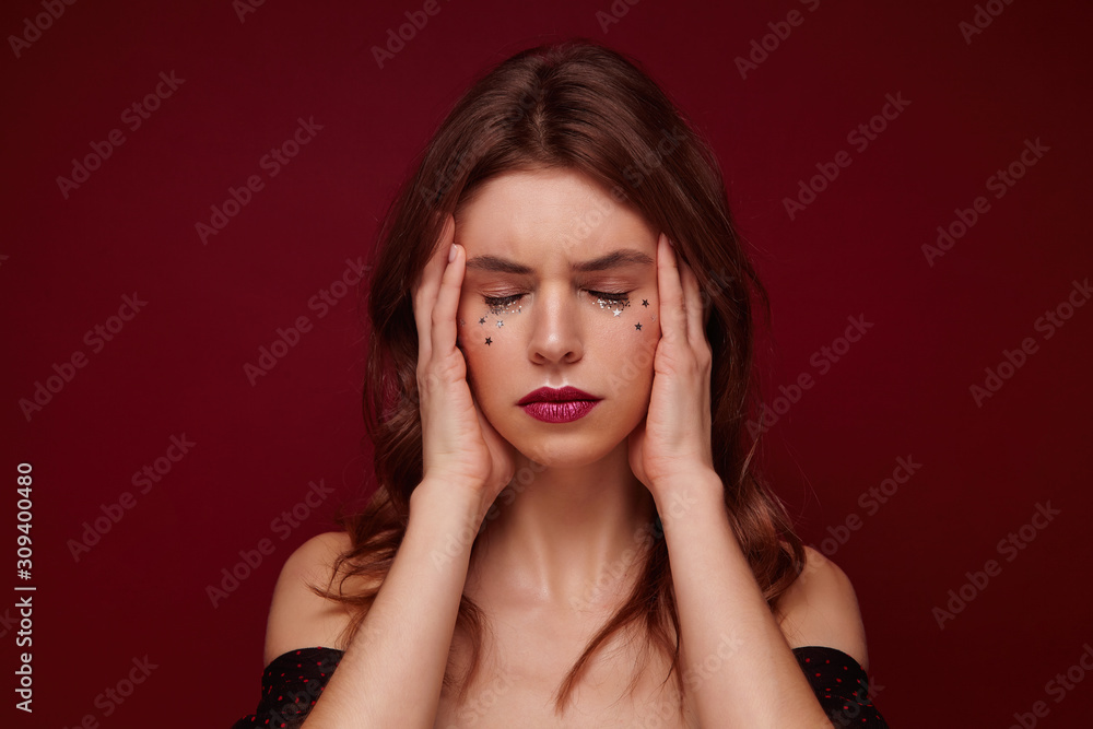 Stressed young brunette lady with curls looking tired and holding her face with hands, keeping eyes closed and frowning eyebrows while posing over claret background Stressed young brunette lady with curls looking tired and holding her face with hands, keeping eyes closed and frowning eyebrows while posing over claret background