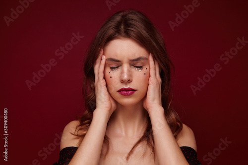 Stressed young brunette lady with curls looking tired and holding her face with hands, keeping eyes closed and frowning eyebrows while posing over claret background