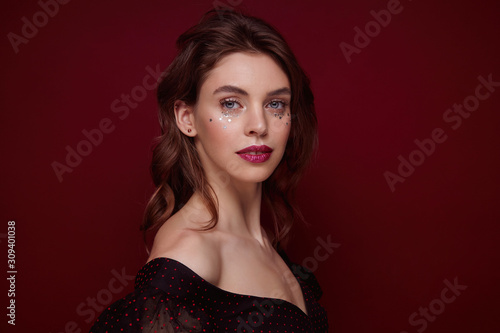Portrait of charming young brunette lady with wavy hairstyle wearing evening makeup while posing over claret background, dressed in festive clothes with deep neckline photo