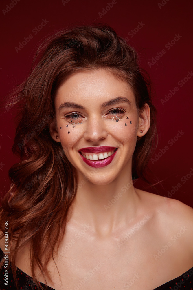 Fototapeta premium Close-up of young pretty brunette lady with evening makeup looking happily at camera and smiling widely, being in high spirit while posing against claret background