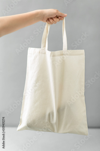 consumerism and eco friendly concept - hand holding reusable canvas bag for food shopping on grey background