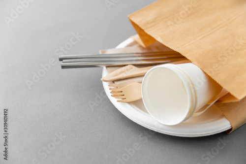 recycling and eco friendly concept - set of wooden forks, knives, paper cups and metallic straws with napkin on plate on grey background photo