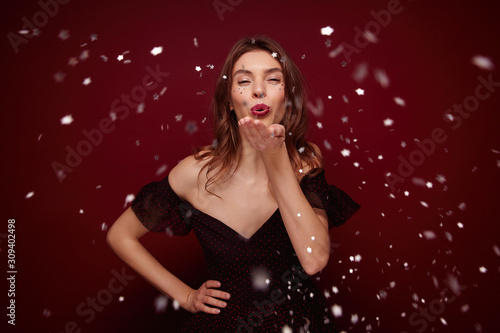 Studio photo of young brunette female dressed in elegant dress enjoying new year theme party while posing over claret background, raising palm and blowing silver confetti to camera