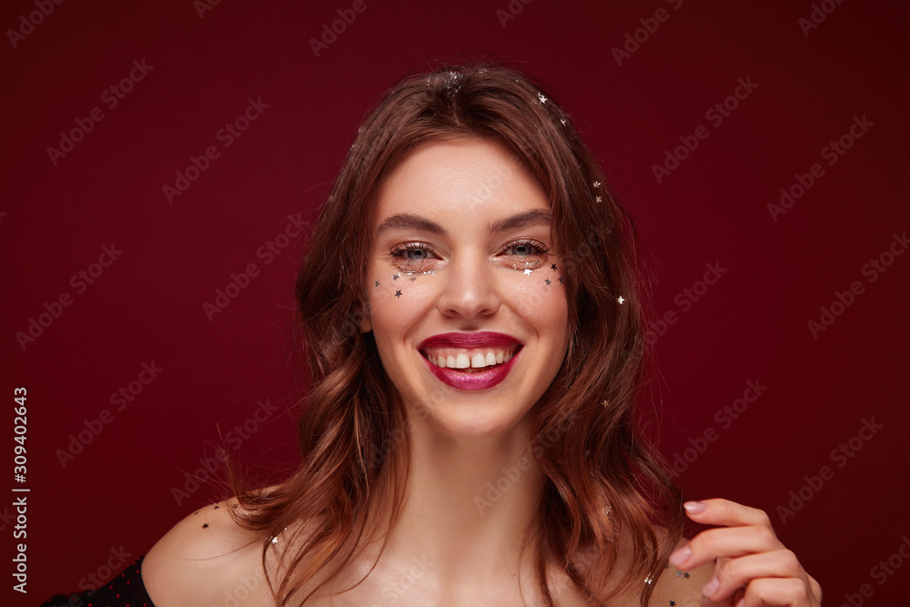 Portrait of lovely cheerful young brown haired female with festive makeup and silver stars on her face preparing for new year party, smiling widely to camera over claret background Portrait of lovely cheerful young brown haired female with festive makeup and silver stars on her face preparing for new year party, smiling widely to camera over claret background
