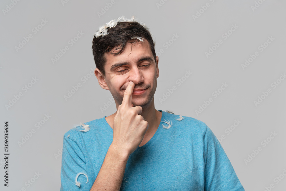 Young man with white feather from a pillow over grey background. Tired after long day Young man with white feather from a pillow over grey background. Tired after long day