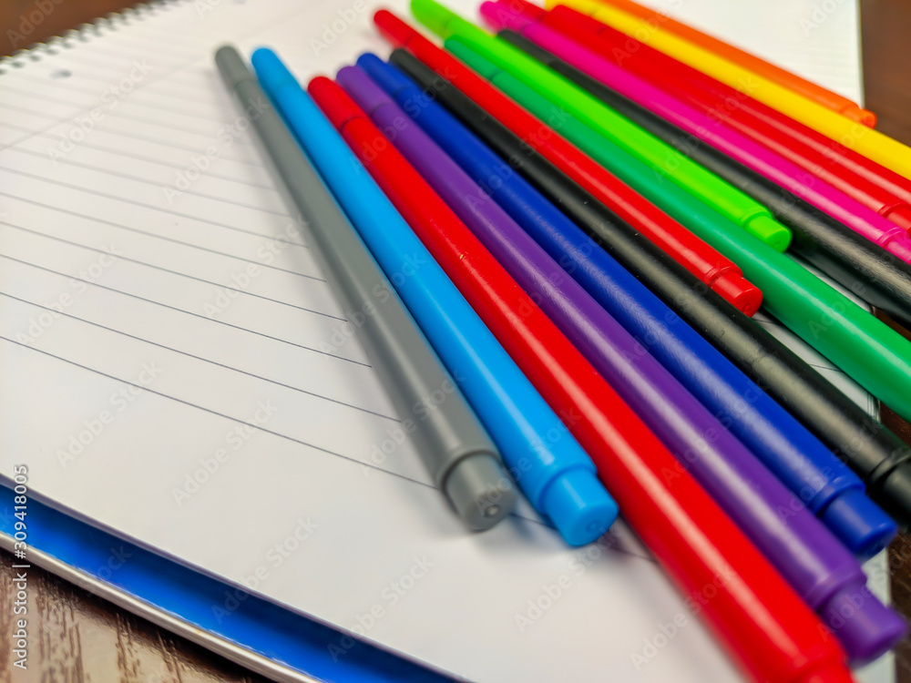 Closeup of colorful pens laying on an empty white notebook for writing down notes as an equipment or supplies for school or studying Closeup of colorful pens laying on an empty white notebook for writing down notes as an equipment or supplies for school or studying