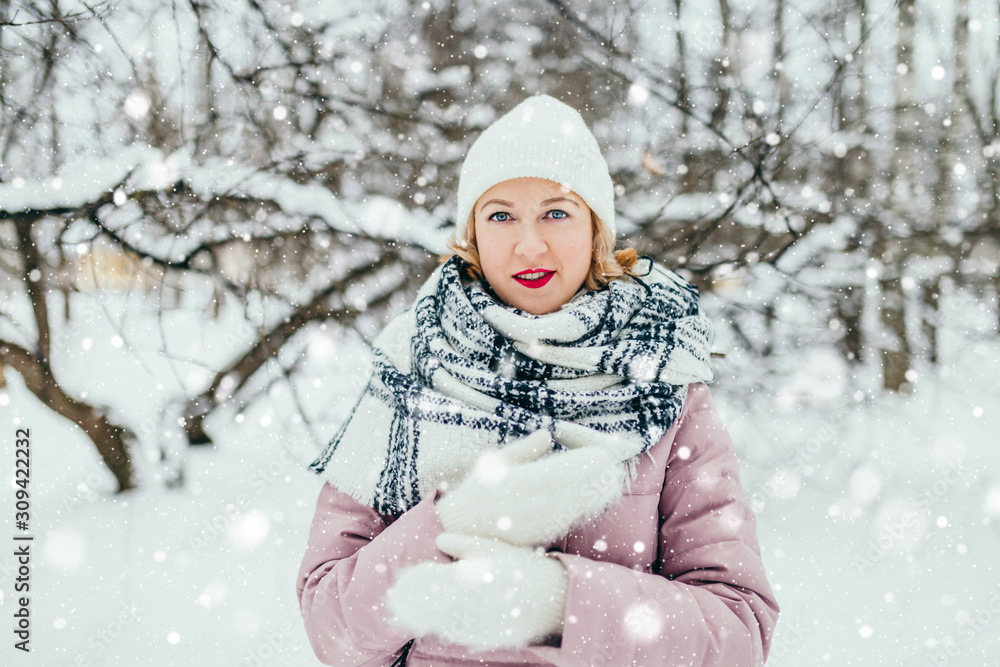 Fototapeta premium Young attractive blonde in a warm down jacket, knitted mittens, a scarf and a hat in a cold winter forest - warm and cozy clothes