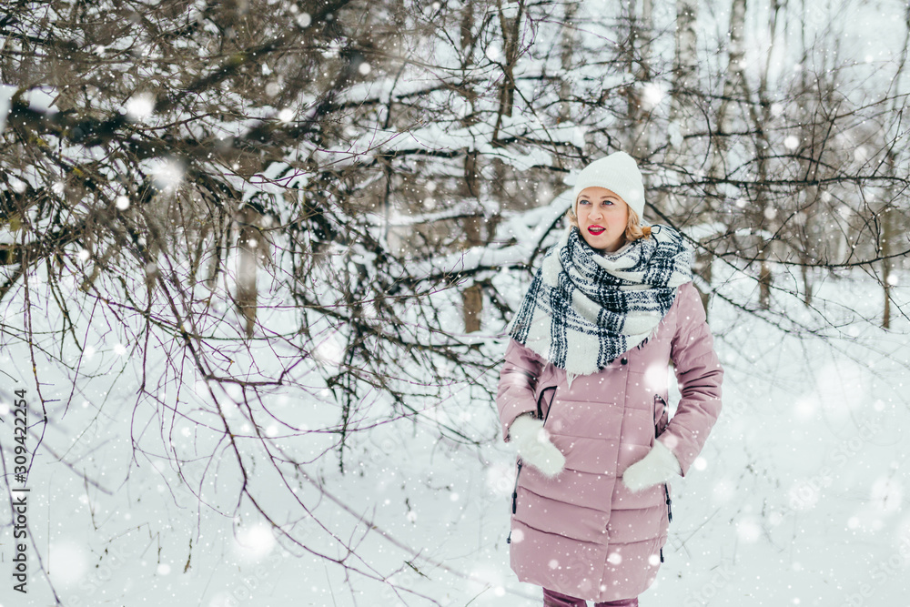 Fototapeta premium Young attractive blonde in a warm down jacket, knitted mittens, a scarf and a hat in a cold winter forest - warm and cozy clothes