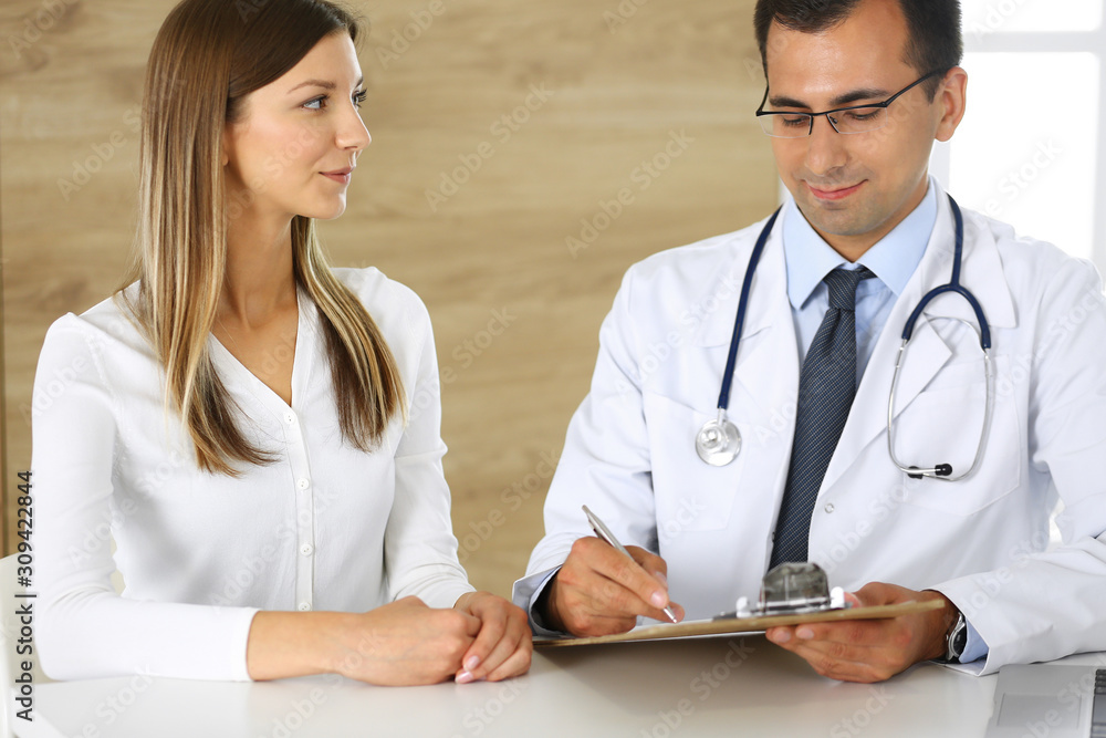 Doctor and patient discussing the results of a physical examination while sitting at a desk in a clinic. A male doctor using a clipboard to fill out a medical history of a young woman's medication Doctor and patient discussing the results of a physical examination while sitting at a desk in a clinic. A male doctor using a clipboard to fill out a medical history of a young woman's medication