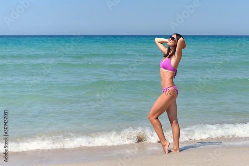 A young woman in a lilac bikini stands on a sandy beach and sunbathes under the bright sun in the hot summer