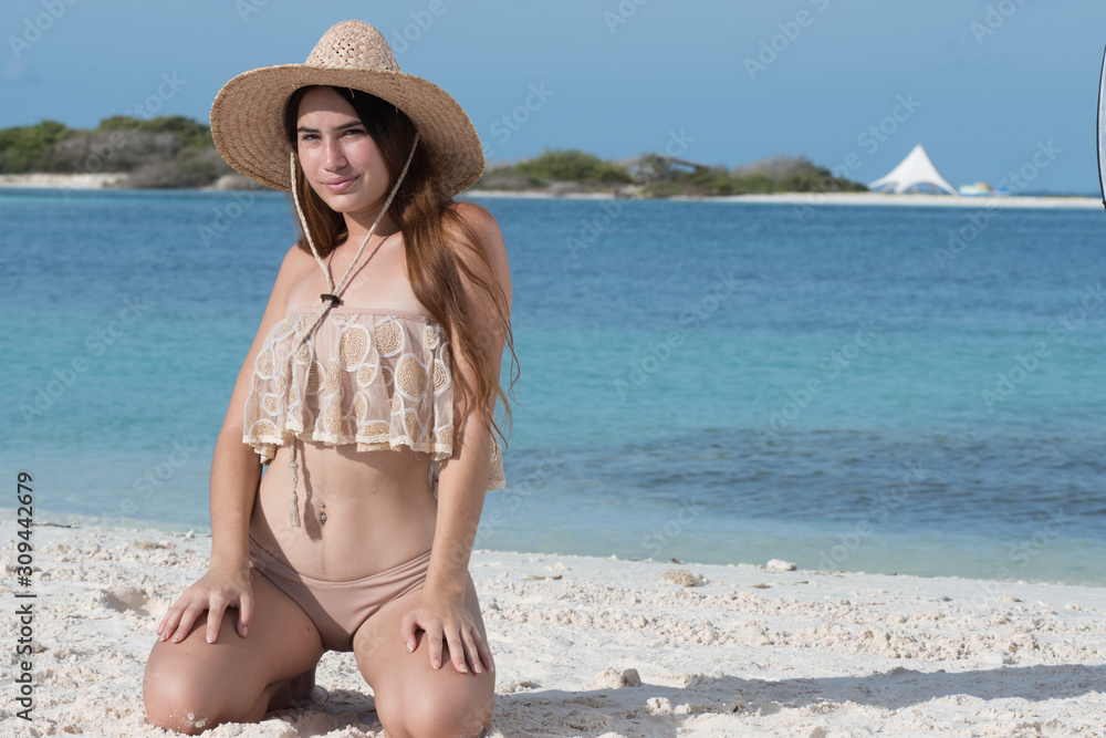 Young caucasian tanned girl kneeling on sand in swimwear looking at camera with blue water beach background