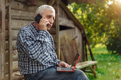 Elderly man with a laptop in village, in nature. Old man out of town with computer. Grandfather in country communicates with his family via the Internet